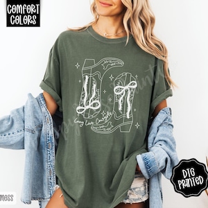 May include: Moss green t-shirt with a white graphic of two cowboy boots and the text "Long Live Cowgirls". The shirt is a Comfort Colors brand and has a relaxed fit. The design is DTG printed.