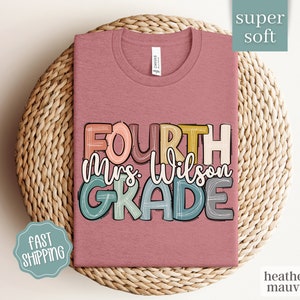 May include: Heather mauve t-shirt with the text "FOURTH Mrs. Wilson GRADE" in colorful, outlined letters. The shirt is displayed on a woven basket.