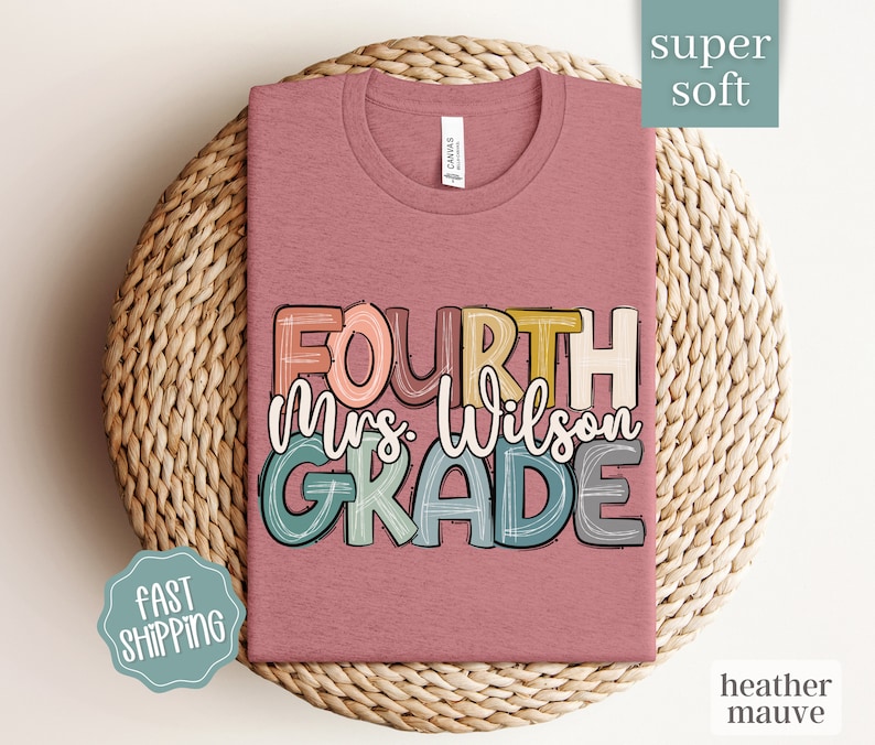 May include: Heather mauve t-shirt with the text "FOURTH Mrs. Wilson GRADE" in colorful, outlined letters. The shirt is displayed on a woven basket.