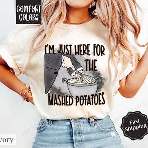 May include: A white t-shirt with a graphic of a hand stirring mashed potatoes in a bowl. The text "I'm just here for the mashed potatoes" is printed on the shirt.