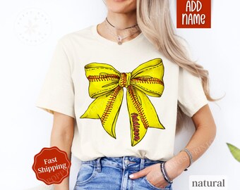 May include: A white t-shirt featuring a yellow softball bow with red stitching. The bow has the name "Addison" written on it.
