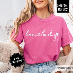 May include: A pink t-shirt with the text "homebody" in white cursive lettering and a small white heart. The shirt is made of a soft, comfortable material.