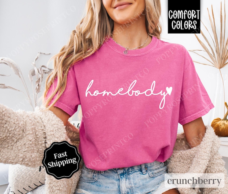 May include: A pink t-shirt with the text "homebody" in white cursive lettering and a small white heart. The shirt is made of a soft, comfortable material.