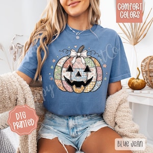 May include: A blue t-shirt featuring a patchwork pumpkin design with a smiling face, bow, and floral accents. The shirt has the text "Comfort Colors" and "Blue Jean". The design is printed on the shirt.