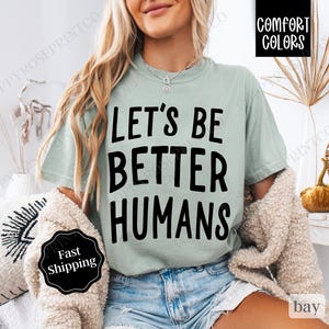 May include: A light green t-shirt with the text "Let's Be Better Humans" printed in black. The shirt is being worn by a person.