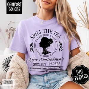 Pu&ograve; includere: Una maglietta lilla con la scritta "SPILL THE TEA" e "Lady Whistledown's SOCIETY PAPERS" in nero. Il design include la silhouette di una donna in un cerchio. La maglietta presenta anche le parole "COMFORT COLORS" e "DTG PRINTED".