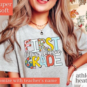 May include: A grey t-shirt with the text "FIRST Mrs. Wilson GRADE" in colourful, patterned letters. The shirt is made of a soft, athletic heather fabric.