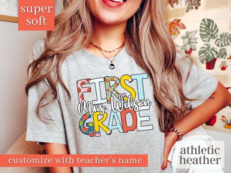 May include: A grey t-shirt with the text "FIRST Mrs. Wilson GRADE" in colourful, patterned letters. The shirt is made of a soft, athletic heather fabric.