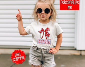 May include: White t-shirt with a red, white, and blue baseball graphic and a bow. The text "Sophia" is on a blue and white checkered banner below the baseball.