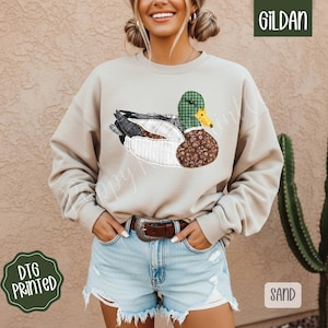 May include: A sand-colored sweatshirt featuring a detailed duck design. The duck is composed of various patterned fabrics, including plaid, floral, and textured materials. The sweatshirt has a crew neck and long sleeves. The image also includes the text "DTG PRINTED" and "SAND".
