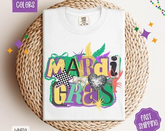 May include: White t-shirt with a colorful "Mardi Gras" graphic. The design includes a checkered star, a sequined heart, a bow, and a crawfish. The shirt is displayed on a woven surface, with the words "Comfort Colors" and "White" visible.