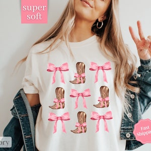 May include: White t-shirt with a pink bow and brown cowboy boot design. The text "super soft" is in a pink box at the top of the image. The text "ivory" is in a pink box at the bottom of the image.