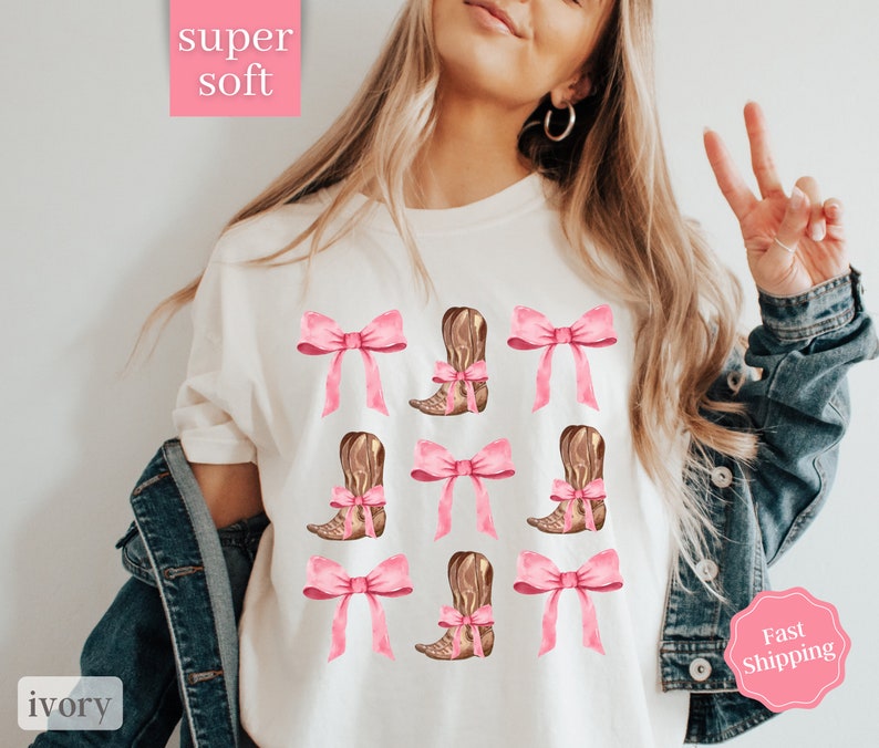 May include: White t-shirt with a pink bow and brown cowboy boot design. The text "super soft" is in a pink box at the top of the image. The text "ivory" is in a pink box at the bottom of the image.