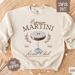 May include: A beige sweatshirt with the text "Espresso Martini" in brown lettering. The sweatshirt also features a graphic of a martini glass with coffee and a brown leaf design. The text "Tini Time" is below the glass and the text "Social Club" is below that. The text "Vodka, Coffee Liqueur, Espresso, Simple Syrup" is below the "Social Club" text.