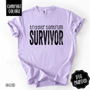 May include: A light purple t-shirt with the words "toddler tantrum SURVIVOR" printed in black. The shirt has short sleeves and a crew neck. The text "COMFORT COLORS" is in a black box in the upper left corner. The text "DTG PRINTED" is in a black circle in the lower right corner.