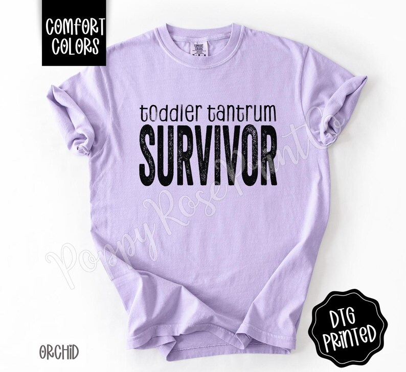 May include: A light purple t-shirt with the words "toddler tantrum SURVIVOR" printed in black. The shirt has short sleeves and a crew neck. The text "COMFORT COLORS" is in a black box in the upper left corner. The text "DTG PRINTED" is in a black circle in the lower right corner.