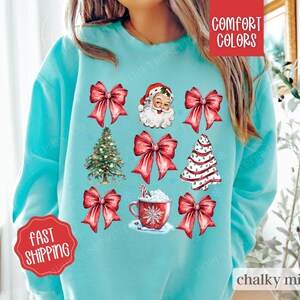 May include: A turquoise blue sweatshirt with a Christmas theme featuring Santa Claus, Christmas trees, bows, and a cup of hot chocolate. The text "COMFORT COLORS" is in the top right corner and "chalky mint" is in the bottom right corner.