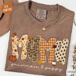 May include: A brown short-sleeve t-shirt featuring the word MAMA in large letters with varied patterns including floral, leopard print, orange gingham, and brown polka dots. The design includes small autumn leaves and a pumpkin graphic.
