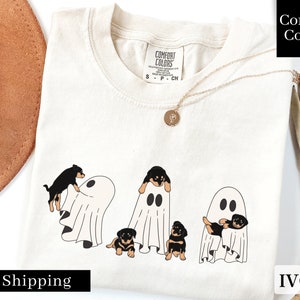 May include: A white t-shirt with a graphic of four black and brown puppies peeking out from behind white ghosts. The text "Comfort Colors" is in the top right corner of the image. The text "IVORY" is in the bottom right corner of the image. The text "Fast Shipping" is in the bottom left corner of the image.