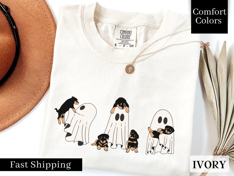 May include: A white t-shirt with a graphic of four black and brown puppies peeking out from behind white ghosts. The text "Comfort Colors" is in the top right corner of the image. The text "IVORY" is in the bottom right corner of the image. The text "Fast Shipping" is in the bottom left corner of the image.