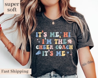 May include: A grey t-shirt with the text "It's me, Hi I'm the Cheer Coach It's me" in colourful letters with a star and a cheerleader pom-pom graphic.