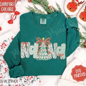 Puede incluir: Una sudadera de manga larga color verde azulado con la palabra "Noel" en un diseño festivo. La "N" y la "d" están decoradas con lazos y patrones, y la "oel" presenta un árbol de Navidad. También son visibles las palabras "Comfort Colors" y "Blue Spruce".