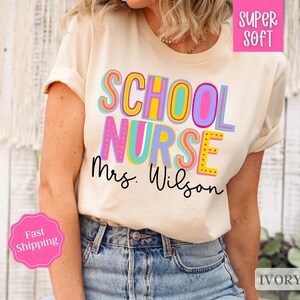 Puede incluir: Una camiseta blanca con el texto "School Nurse Mrs. Wilson" en letras de colores. La camiseta es s&uacute;per suave y de color marfil.