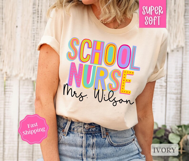Puede incluir: Una camiseta blanca con el texto "School Nurse Mrs. Wilson" en letras de colores. La camiseta es s&uacute;per suave y de color marfil.