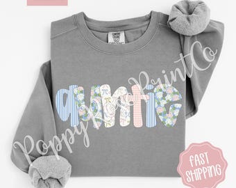 May include: A grey sweatshirt with the word "Auntie" in a patchwork design. The letters are decorated with blue stripes, floral patterns, and pink gingham. The sweatshirt has a scrunchie on the shoulder and the words "Comfort Colors" are in the top left corner.