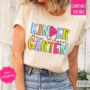 May include: A cream colored t-shirt with the text "Kinder" in rainbow colors and "Mrs. Wilson Kindergarten" in colorful letters with a variety of school-themed illustrations.