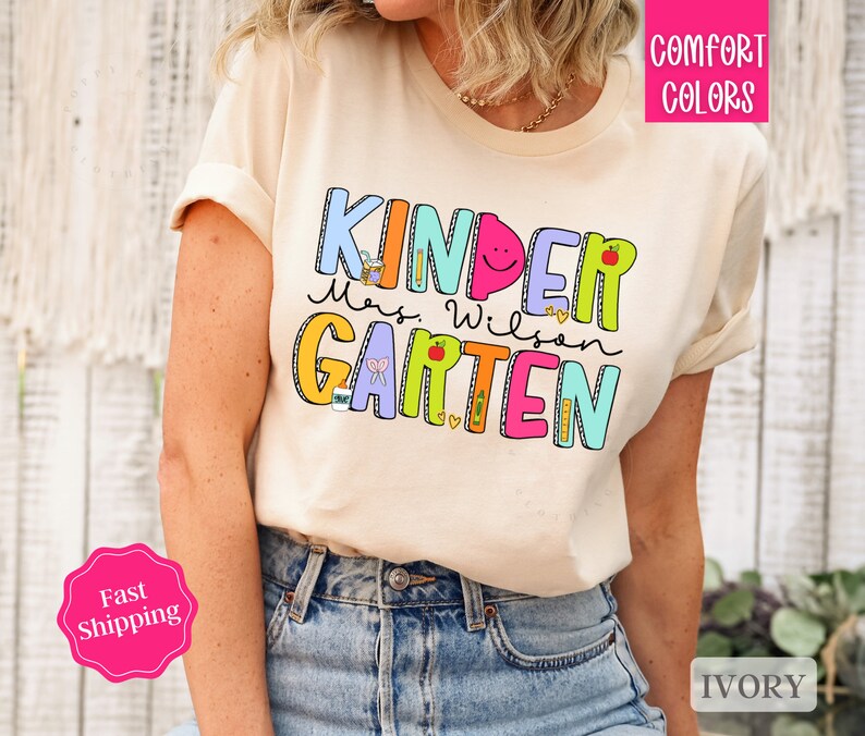 May include: A cream colored t-shirt with the text "Kinder" in rainbow colors and "Mrs. Wilson Kindergarten" in colorful letters with a variety of school-themed illustrations.