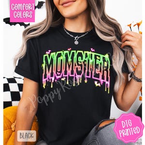 May include: Black t-shirt with the word "MOMSTER" in neon green and pink, with dripping accents and bat graphics. The shirt is labeled "Comfort Colors" and "DTG Printed".