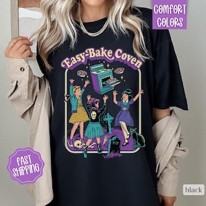 May include: A black t-shirt featuring a graphic print with the text "Easy-Bake Coven". The design depicts three figures in a graveyard setting with a purple oven, a tombstone marked "RIP", a black cat, and a skull, all in a retro comic style.