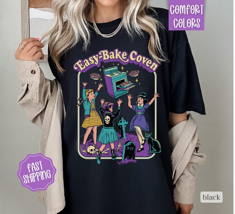 May include: A black t-shirt featuring a graphic print with the text "Easy-Bake Coven". The design depicts three figures in a graveyard setting with a purple oven, a tombstone marked "RIP", a black cat, and a skull, all in a retro comic style.
