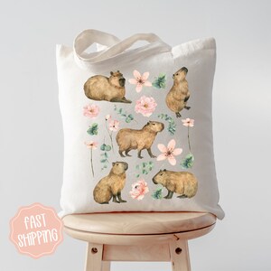 Pu&ograve; includere: Una borsa tote in tela bianca con un design floreale con fiori rosa e foglie verdi. La borsa ha un'illustrazione di capibara marrone su ogni lato.