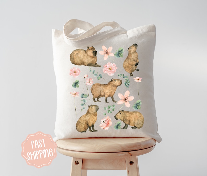 Pu&ograve; includere: Una borsa tote in tela bianca con un design floreale con fiori rosa e foglie verdi. La borsa ha un'illustrazione di capibara marrone su ogni lato.