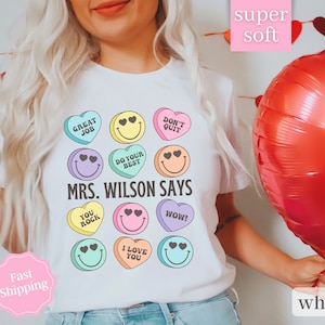 Puede incluir: Camiseta blanca con un gráfico de corazones de caramelo coloridos con lemas como "Great Job", "Do Your Best", "You Rock" e "I Love You". El texto "Mrs. Wilson Says" está impreso debajo de los corazones.