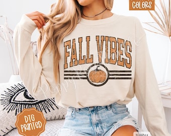 May include: Cream long-sleeved top with the words "FALL VIBES" in orange and brown, and a pumpkin graphic. The shirt also has the text "COMFORT COLORS" and "DTG PRINTED".