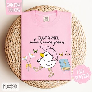 Puede incluir: Camiseta rosa con un pato de dibujos animados que lleva botas de lluvia y sostiene un paraguas. La camiseta tiene el texto "Just a girl who loves Jesus" y una imagen de una biblia.