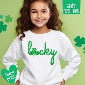 May include: White sweatshirt with a green glitter 'lucky' design featuring a shamrock. The sweatshirt has a crew neck and long sleeves.