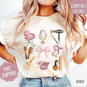 May include: A white t-shirt with a design of hair salon tools, including a hair dryer, scissors, combs, a hair dye bowl, and a hair tie. The text "Comfort Colors" is in the top right corner of the image. The t-shirt is ivory.