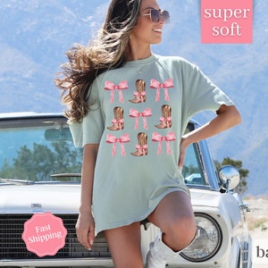 May include: A light blue t-shirt with a graphic print of pink bows and brown cowboy boots. The text "super soft" is printed in white on a pink background.