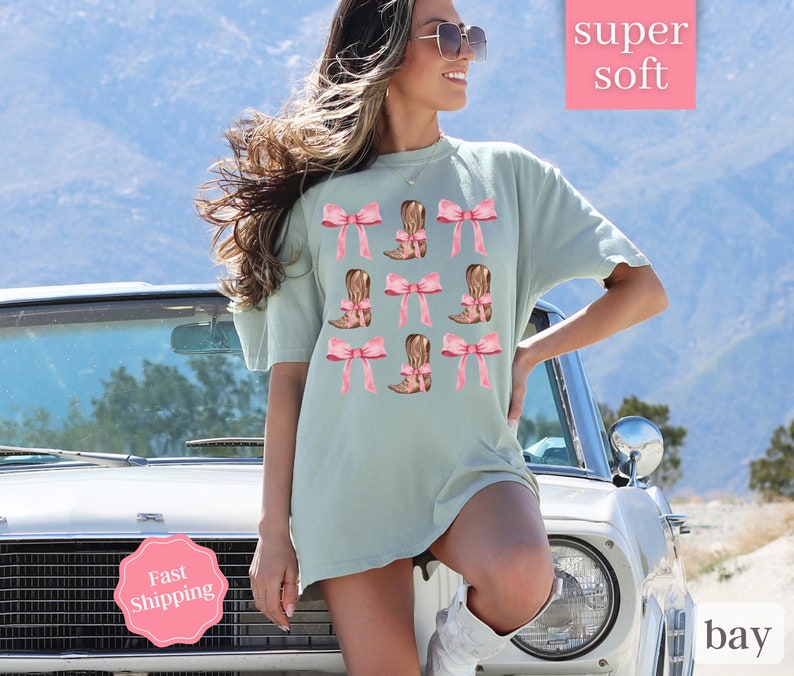 May include: A light blue t-shirt with a graphic print of pink bows and brown cowboy boots. The text "super soft" is printed in white on a pink background.