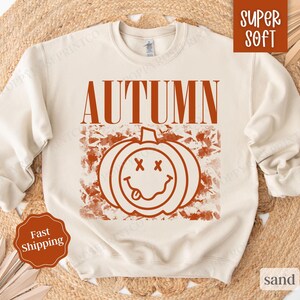 May include: A cream colored sweatshirt with the word "AUTUMN" printed in orange on the front. The word is above a graphic of a smiling pumpkin with a tongue sticking out.