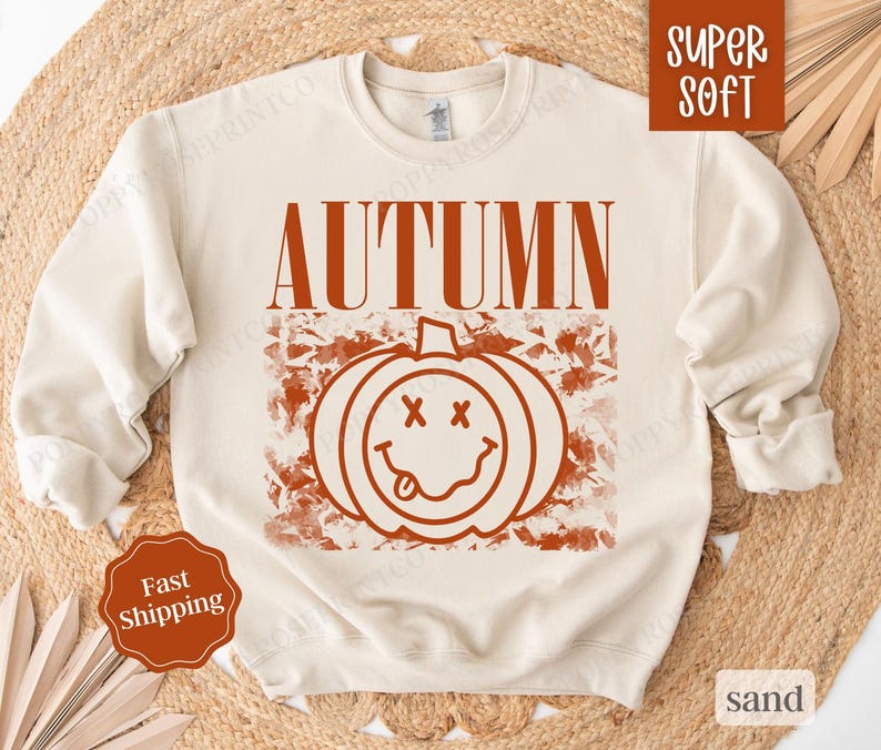 May include: A cream colored sweatshirt with the word "AUTUMN" printed in orange on the front. The word is above a graphic of a smiling pumpkin with a tongue sticking out.