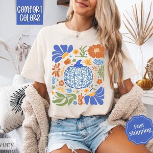 Op de afbeelding: Een wit T-shirt met een blauw en oranje bloemenontwerp met een pompoen met een blauw en wit patroon. De tekst "Comfort Colors" staat bovenaan het shirt gedrukt.