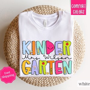 May include: White t-shirt with a colorful graphic that says "Kinder Mrs. Wilson Garten".