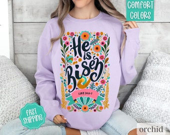 Pode incluir: Uma camisa de moletom lilás com um design floral e o texto "He is risen" com uma referência bíblica de Lucas 24:6-7.