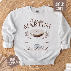 May include: Gray sweatshirt with the text "Espresso MARTINI" and a graphic of a martini glass with coffee beans. The text "TINI TIME Social Club VODKA, COFFEE LIQUEUR, ESPRESSO, SIMPLE SYRUP" is below the graphic.