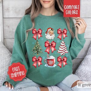 May include: A light green sweatshirt with a Christmas theme featuring Santa Claus, Christmas trees, bows, and a cup of hot chocolate. The sweatshirt has the text "Comfort Colors" on the top right corner and "Fast Shipping" on the bottom left corner.